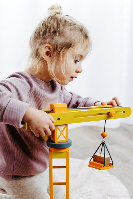 Ein junges Mädchen in einem lila Pullover spielt Rollenspiel und spielt drinnen mit einem gelben Baustellenkran aus Holz.