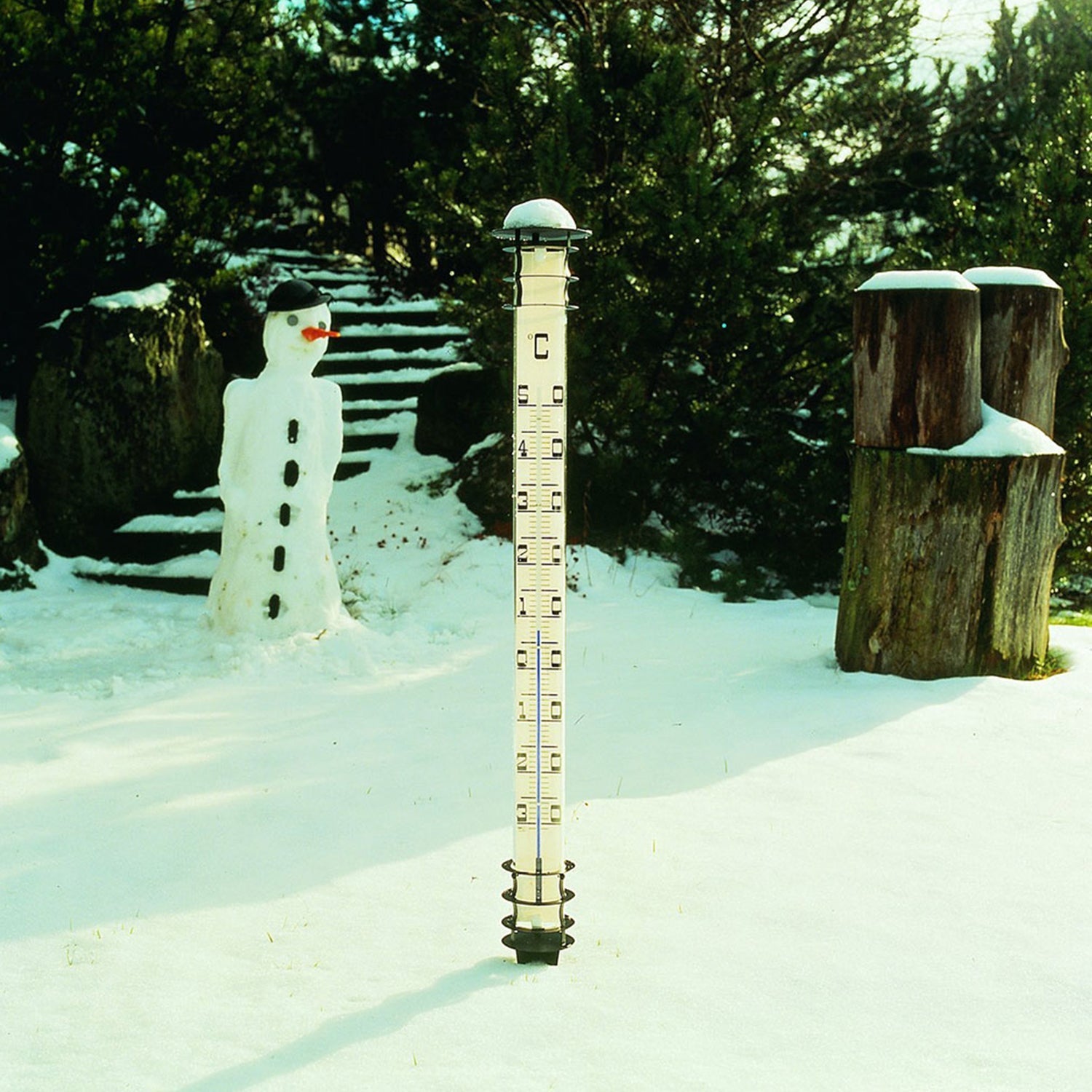 Ein Thermometer für den Garten steht im Schnee neben Schneemann und Bäumen.