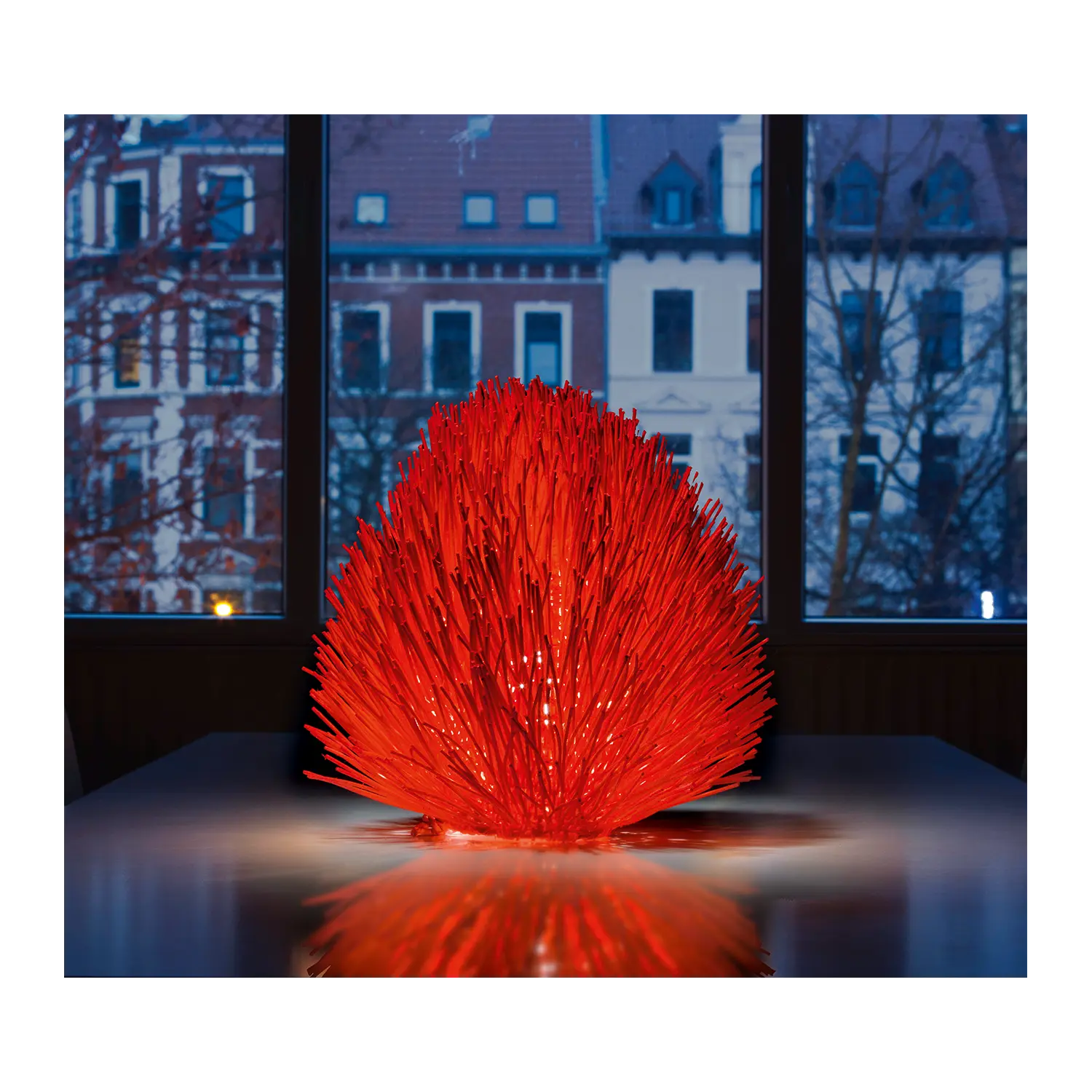 Tischleuchte Trend Rot aus Rattan leuchtet auf einem Tisch an einem Stadtfenster.