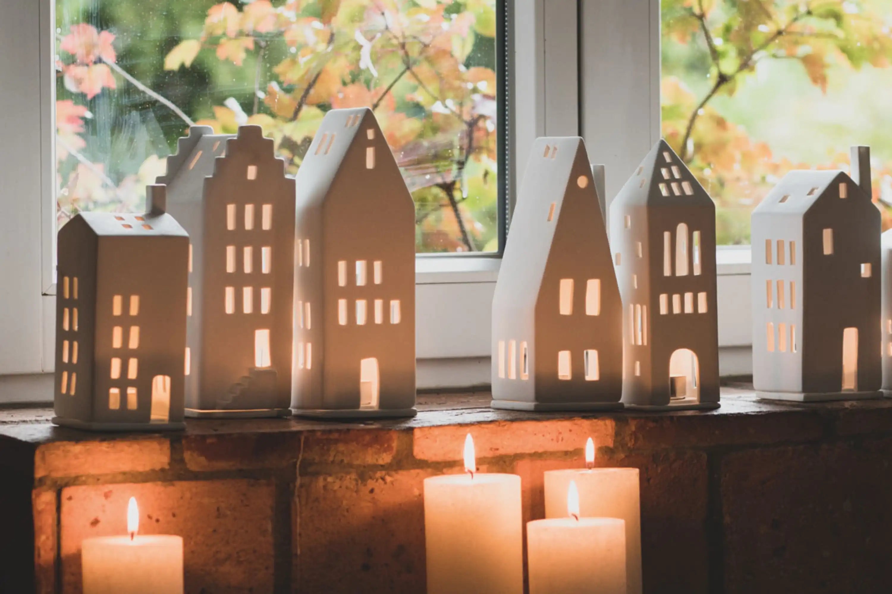 Keramische Hauslaternen und brennende Kerzen auf einer Fensterbank mit Herbstlaub vor dem Fenster.