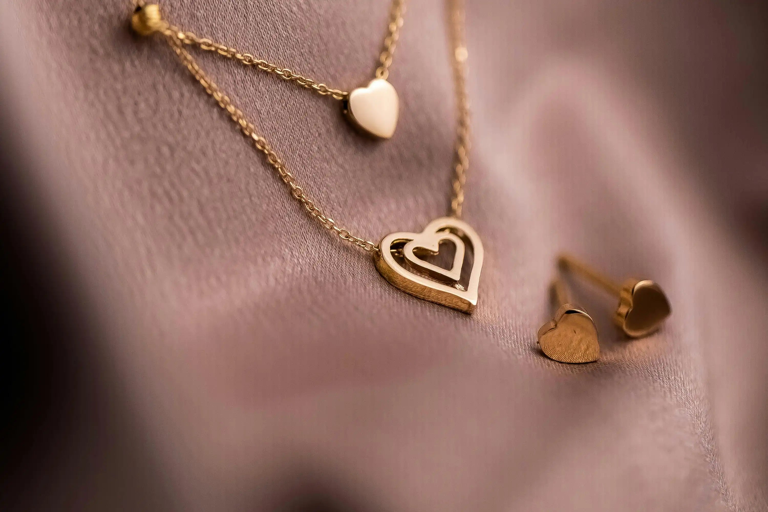 Goldfarbene herzförmige Halskette und Ohrringe auf weichem, rosa Stoff.