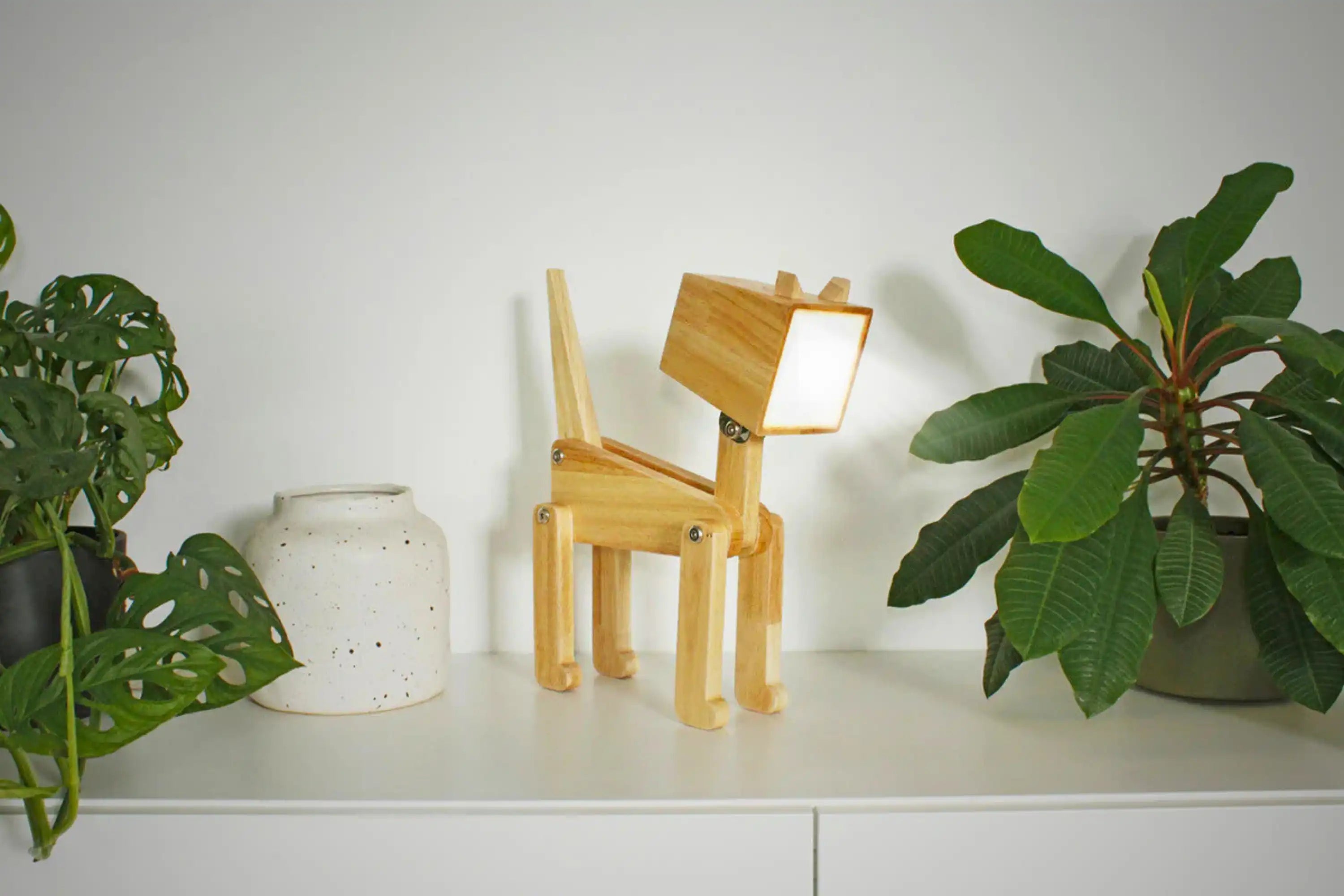 Hölzerne hundeförmige Lampe auf einem weißen Regal zwischen einer Topfpflanze und einer weißen Vase mit grünen Blättern.
