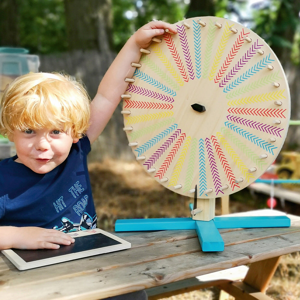 Ein kleines Kind sitzt bei einem Kindergeburtstag im Freien neben einem bunten Glücksrad, auf dem Tisch davor liegt ein Tablet.