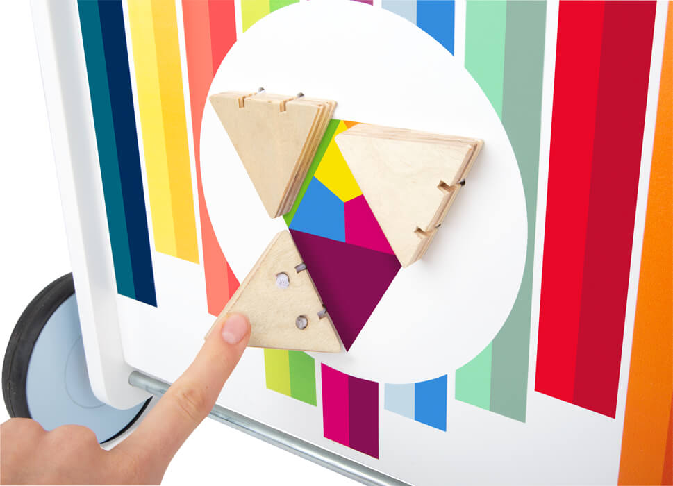 Eine Hand berührt ein dreieckiges Holzpuzzleteil auf einer bunt gestreiften Tafelfläche und fördert so die motorische Entwicklung.