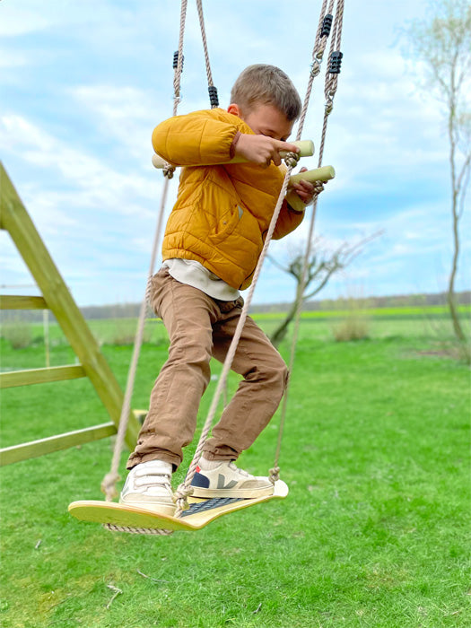 Kleiner Junge in einer gelben Jacke balanciert draußen auf einer Wiese auf einer Skateboard-Schaukel mit verstellbaren Handgriffen.