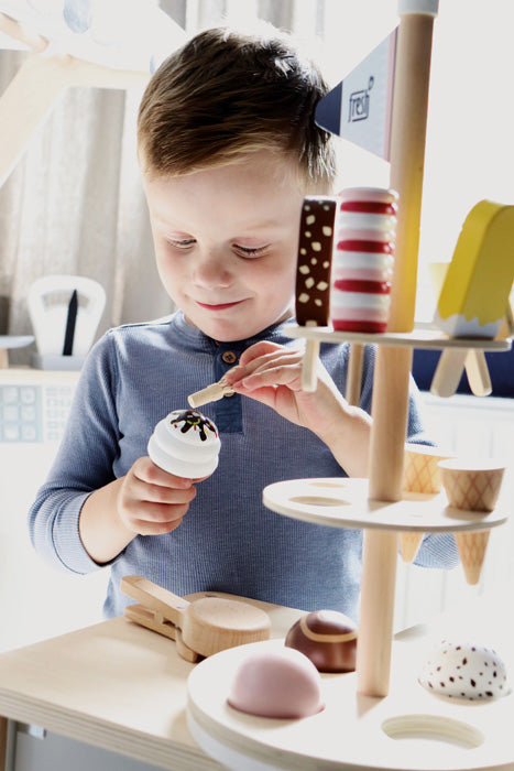 Ein kleiner Junge verwendet einen Eiskugelportionierer, um während einer lustigen Rollenspiel-Eis-Session eine Spielzeug-Eistüte mit Streuseln an einem hölzernen Eisständer aus Holz zu dekorieren.