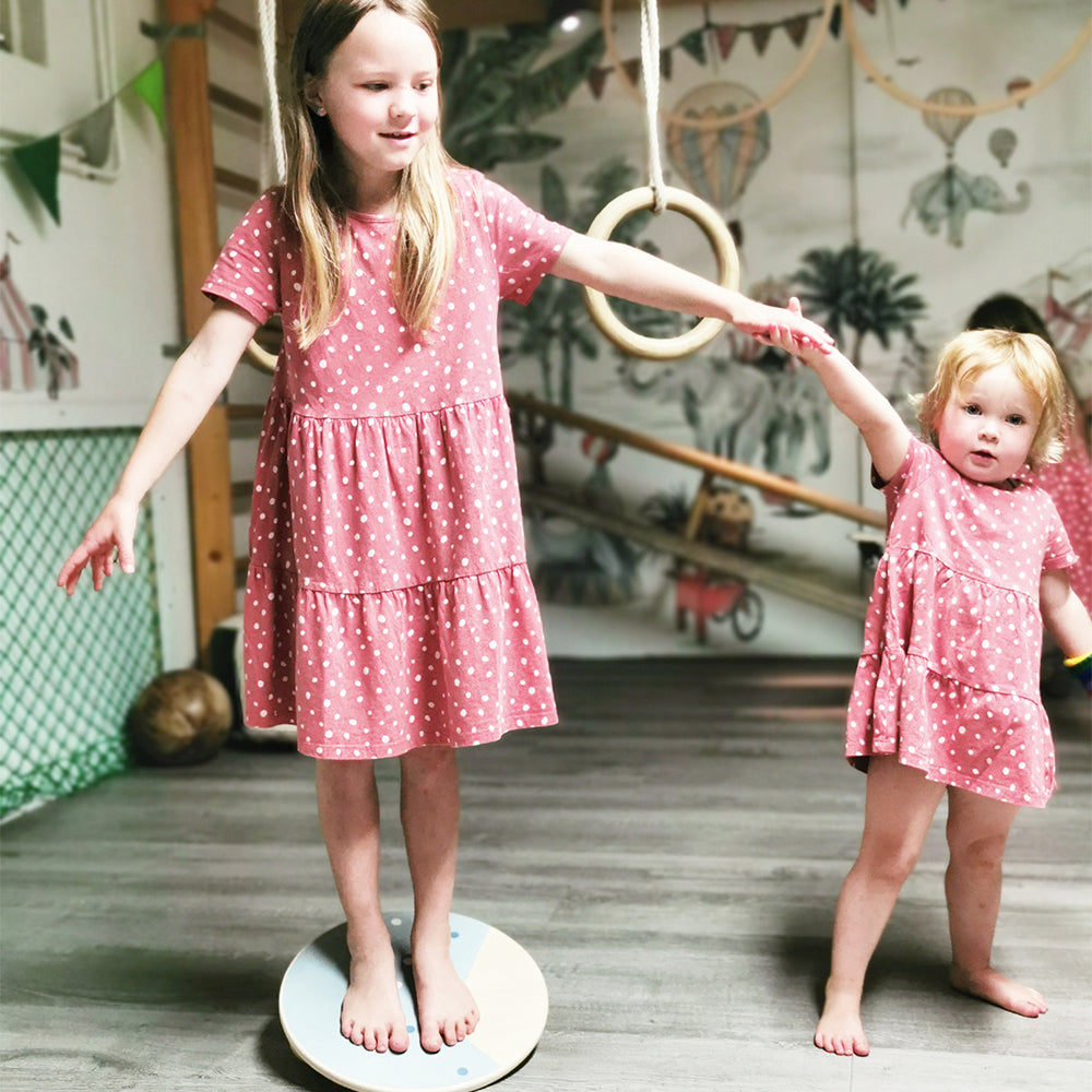 Zwei junge Mädchen in passenden rosa gepunkteten Kleidern balancieren Händchen haltend auf einem Balancierbrett im Innenbereich und genießen gemeinsam ein spielerisches Bewegungstraining.