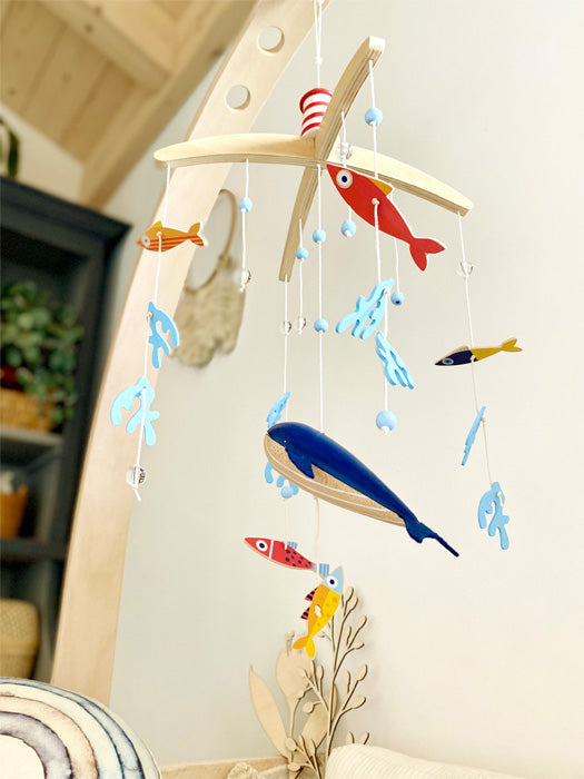 Buntes 3D-Mobile aus Holz mit blauen, roten und gelben Fischen, die an Schnüren hängen und eine verspielte Unterwasserwelt-Atmosphäre in jede Kinderzimmer-Accessoire-Sammlung bringen, im Innenbereich neben einer Pflanze ausgestellt.