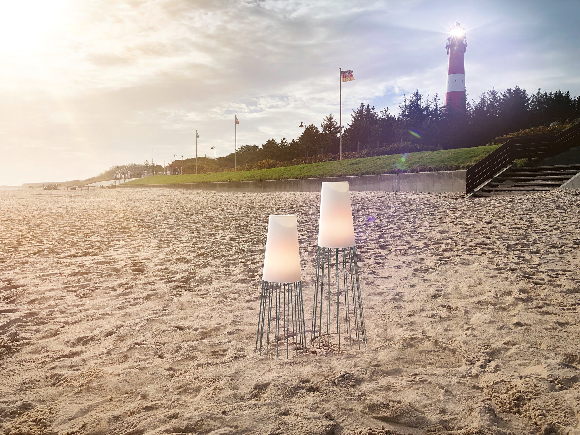 Zwei moderne Lampen verleihen einem Sandstrand bei Sonnenuntergang faszinierende Lichteffekte, wobei der LEUCHTTURM WÄCHTER IN DER NACHT und entfernte Bäume eine ruhige, gartenähnliche Kulisse schaffen.
