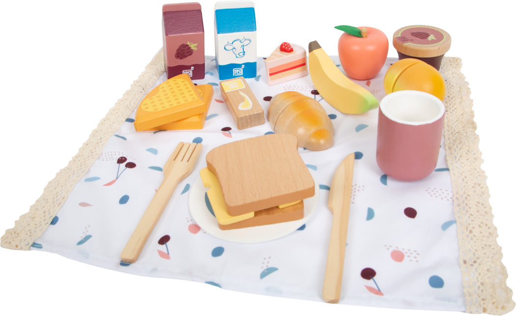 Spielzeug-Frühstücksset aus Holz auf einer gemusterten Picknickdecke, inklusive Brot, Obst, Besteck und Getränken.