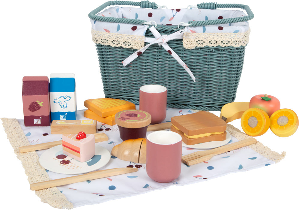Spielzeug-Picknickset mit Picknickkorb, Spielzeuglebensmitteln, Bechern, Tellern und Besteck, hübsch arrangiert auf einer gemusterten Picknickdecke. Dieses bezaubernde Spiel-Set weckt bei Kindern fantasievolle Outdoor-Abenteuer.