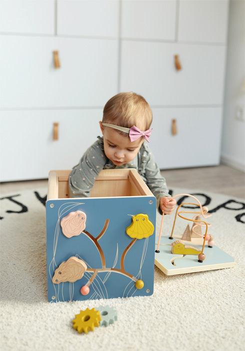 Ein Baby mit einer rosa Schleife spielt mit Holzspielzeug und einem Motorikwürfel auf einem hellen Teppich in einem hellen Raum und erforscht und entwickelt seine Feinmotorik.