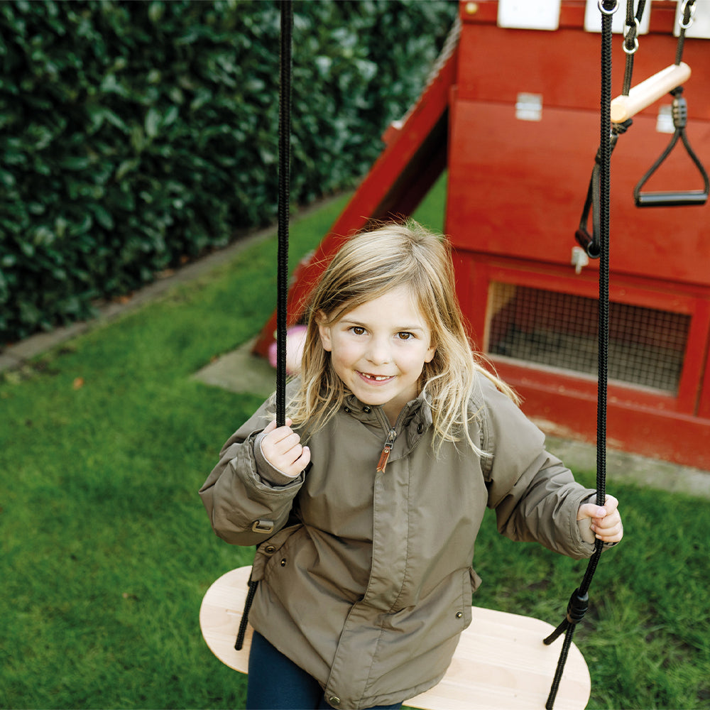 Ein lächelndes Kind in einer grünen Jacke sitzt auf einer Vollholz-Outdoor-Schaukel in einem Hinterhof mit grünem Gras und einem roten Spielhaus.