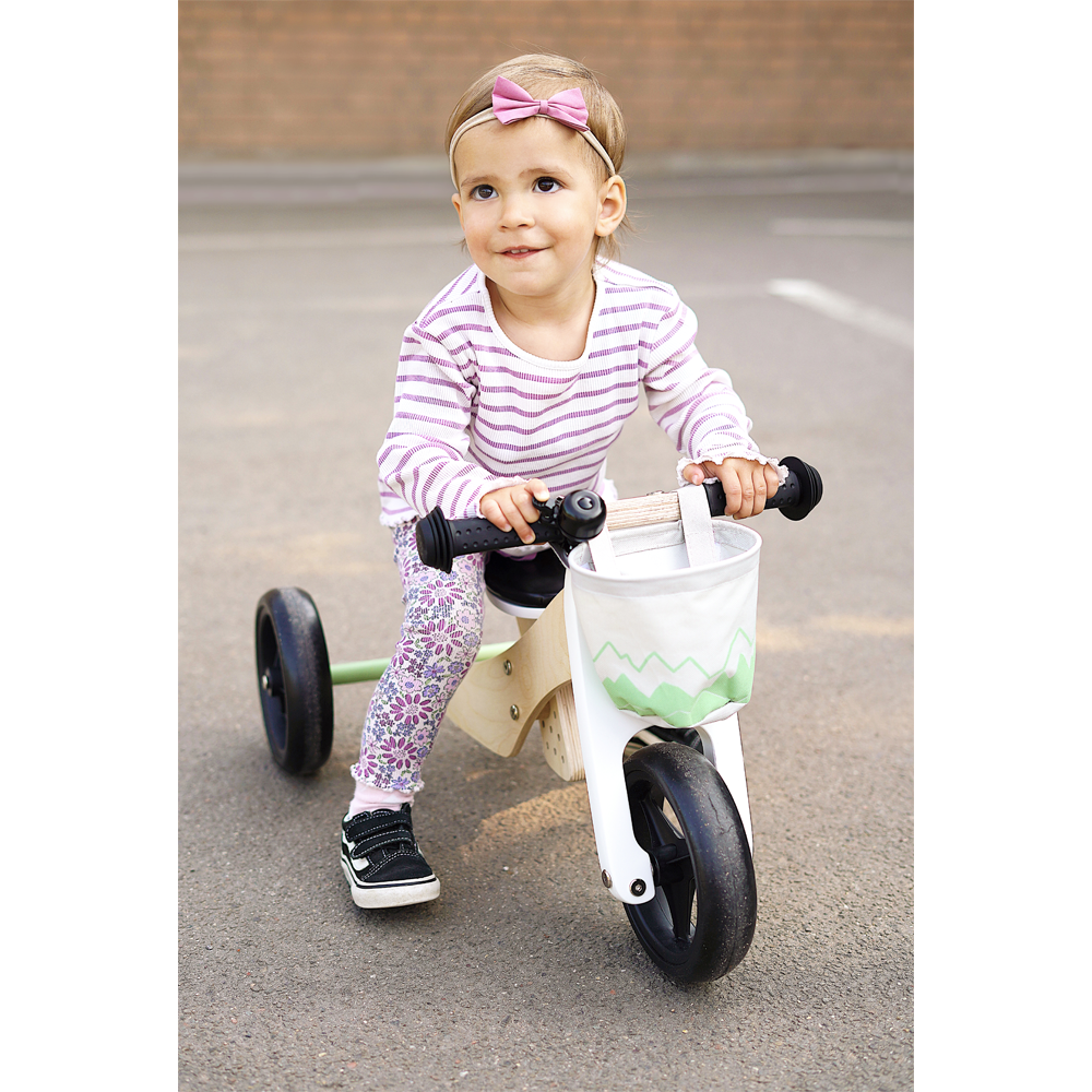 Kleinkind im gestreiften Hemd fährt mit einem Laufrad-Trike über den Bürgersteig, lächelt und trägt ein rosafarbenes Schleifenstirnband – und genießt zusätzliches Gleichgewicht und Sicherheits-Plus.