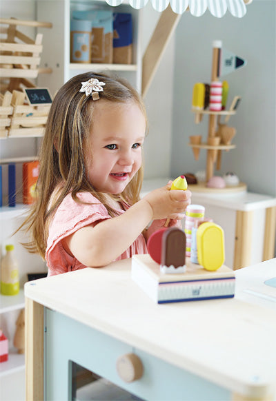 Ein junges Mädchen lächelt, während es in einer fröhlichen Kinderküche mit buntem Spielzeugessen und einem Eisständer spielt.