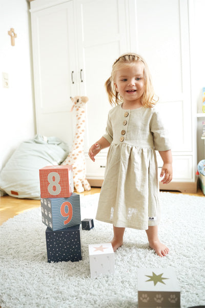 Junges Mädchen im hellen Kleid steht auf einem Teppich neben Stapelwürfeln, Zahlenblöcken mit Polartieren, in einem hellen, aufgeräumten Raum.