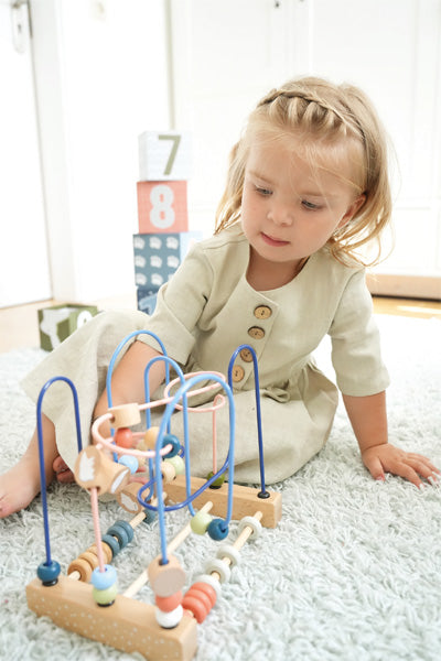Ein junges Mädchen sitzt auf einem Teppich und spielt mit einem Motorikspielzeug aus Holzperlenlabyrinth und verbessert ihre Hand-Augen-Koordination. Im Hintergrund sind Bauklötze zu sehen.