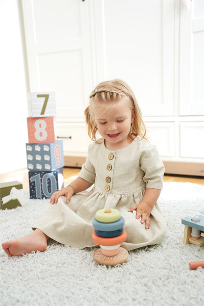 Junges Mädchen in beigem Kleid spielt mit einem bunten Motorikspielzeug-Stapelturm aus FSC-zertifiziertem Holz auf einem weichen weißen Teppich, im Hintergrund Blöcke.