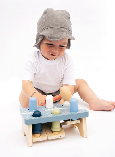 Ein Baby mit grauer Mütze und weißem Hemd spielt mit einem Klopfbank-Holzspielzeug auf dem Boden und verbessert so seine motorischen Fähigkeiten.