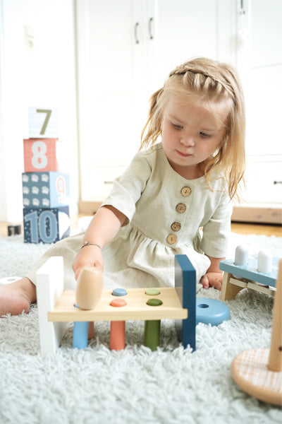 Junges Mädchen in cremefarbenem Kleid spielt mit Hammer und Klopfbank auf einem weichen, hellen Teppich und entwickelt dabei als kleine Motorik-Künstlerin ihre motorischen Fähigkeiten.