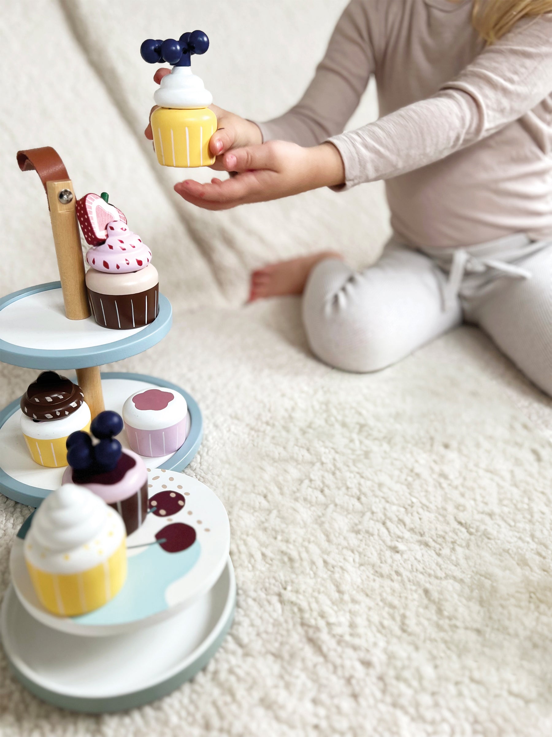 Kind sitzt auf einer Decke und greift nach einem Spielzeug-Cupcake auf einem gestuften Ständer mit Spieldesserts aus Holz aus FSC® 100%-zertifiziertem Holz – perfekt für fantasievolles Rollenspiel rund um die Geburtstagstorte.
