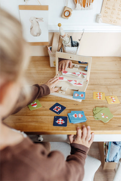 Kind spielt ein Symmetriespiel mit Kartenspiel mit geometrischen Formen und einem Spiegel auf einem Holzschreibtisch in einem gemütlichen, lichtdurchfluteten Raum.