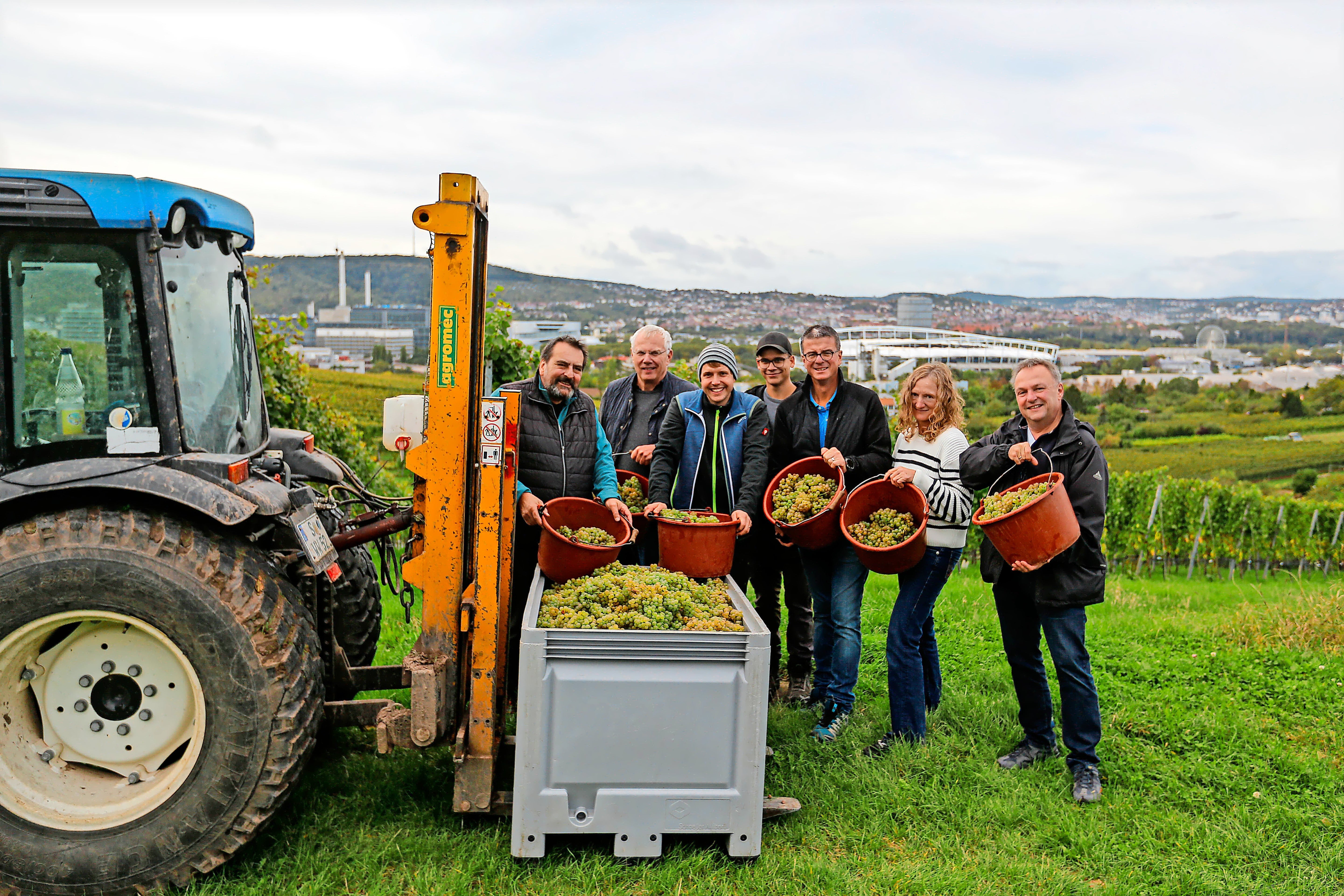 Sieben Menschen stehen lächelnd neben einem Traktor in einem Weinberg des Weinguts Wöhrwag, halten Eimer mit Trauben in den Händen – eine Szene, die den Geist der Stuttgarter Zeitung Weinedition 2025 Merlot + Riesling perfekt einfängt.