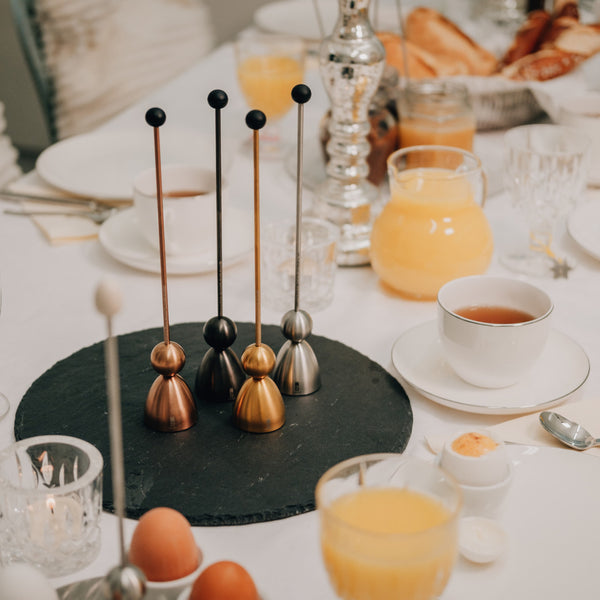 Elegant gedeckter Frühstückstisch mit Saft, Eiern, Tee und dekorativen Edelstahlobjekten auf einem schwarzen runden Tablett, ausgestattet mit einem Eieröffner für den perfekten Eiergenuss.