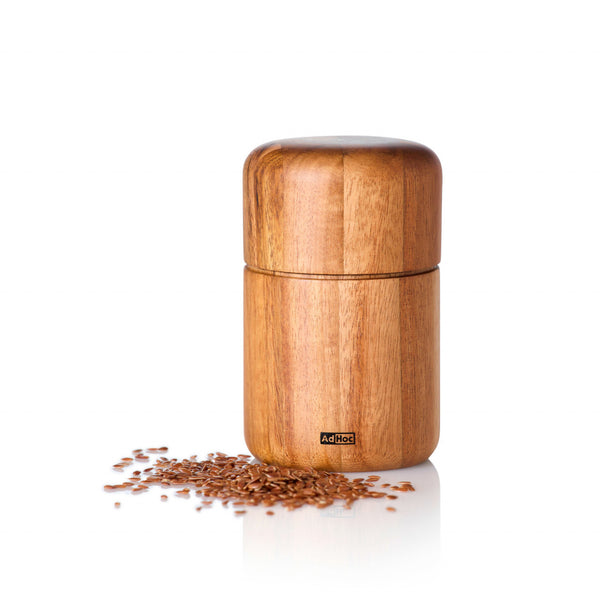 Eine hölzerne Gewürzmühle von Seedo mit einem davor verstreuten Haufen brauner Samen verfügt über das effiziente PolyCut-Mahlwerk und ist somit ideal zum Mahlen selbst ölhaltiger Saaten auf weißem Hintergrund.