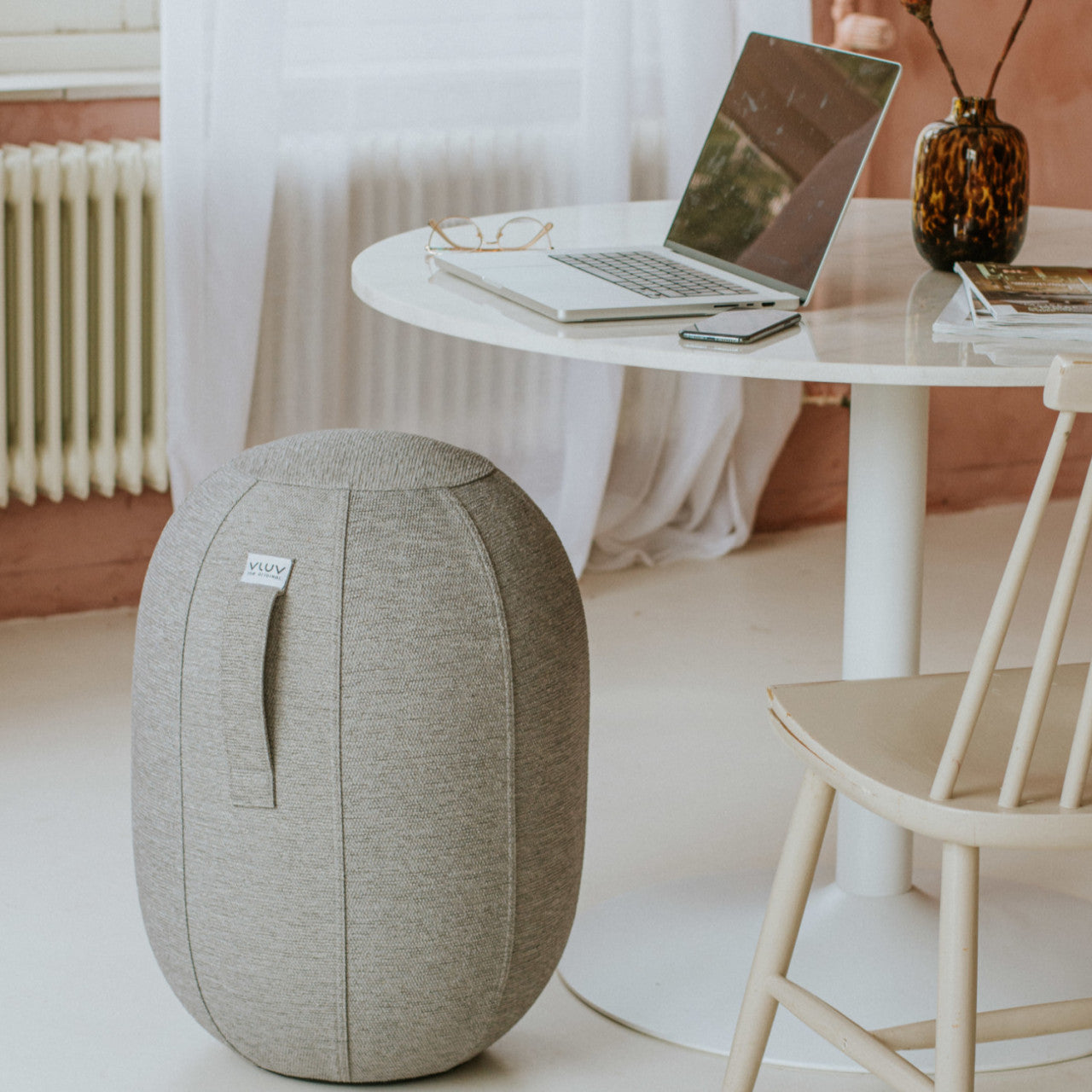 Die graue KAPSUL STOV Balancekapsel sitzt als luftgefüllter Sitzhocker neben einem runden weißen Schreibtisch mit Laptop, Brille und einem Stuhl – perfekt für ergonomisches Sitzen im modernen Büro.