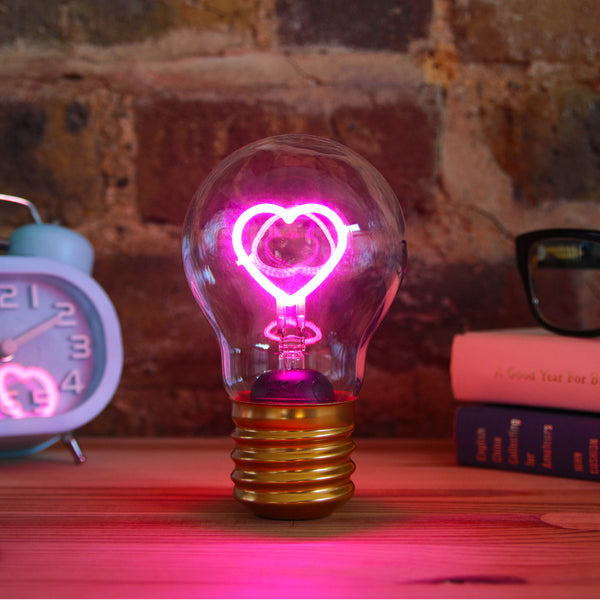 Ein leuchtender rosa Glühfaden in Herzform in einer Edison-Glühbirne, mit Büchern und einer Uhr im Hintergrund.