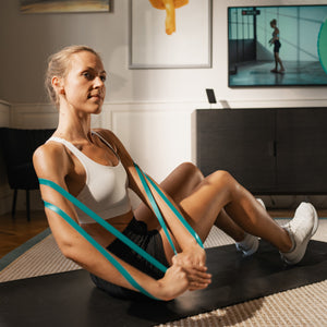 Eine Frau trainiert in ihrem Wohnzimmer auf einer Matte mit Widerstandsbändern und verwendet einen Ganzkörpertrainer für Functional Fitness, während im Hintergrund ein Trainingsvideo im Fernsehen läuft.