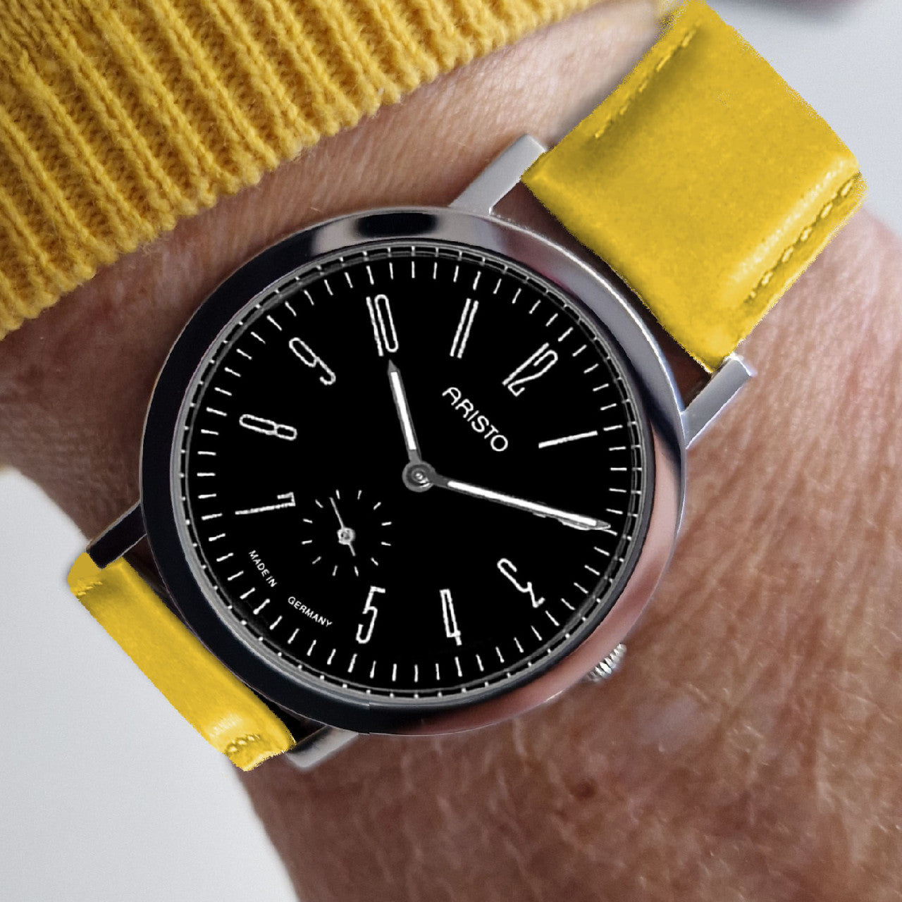 Ein Handgelenk mit einem gelben Uhrenarmband und einer Aristo Bauhaus Uhr mit schwarzem Zifferblatt und Edelstahlgehäuse zeigt die Zeit als 10:08 an.