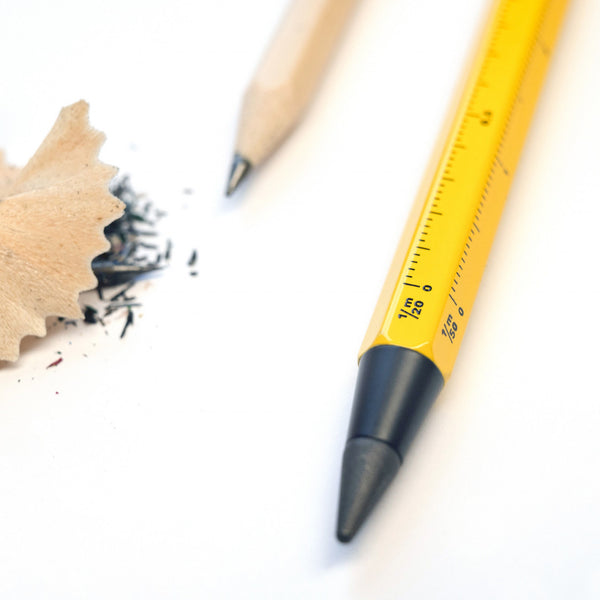 Nahaufnahme eines gelben Multitasking-Bleistifts im BAU-Look, neben einem Holzbleistift, Bleistiftspänen und Graphit auf einer weißen Oberfläche.