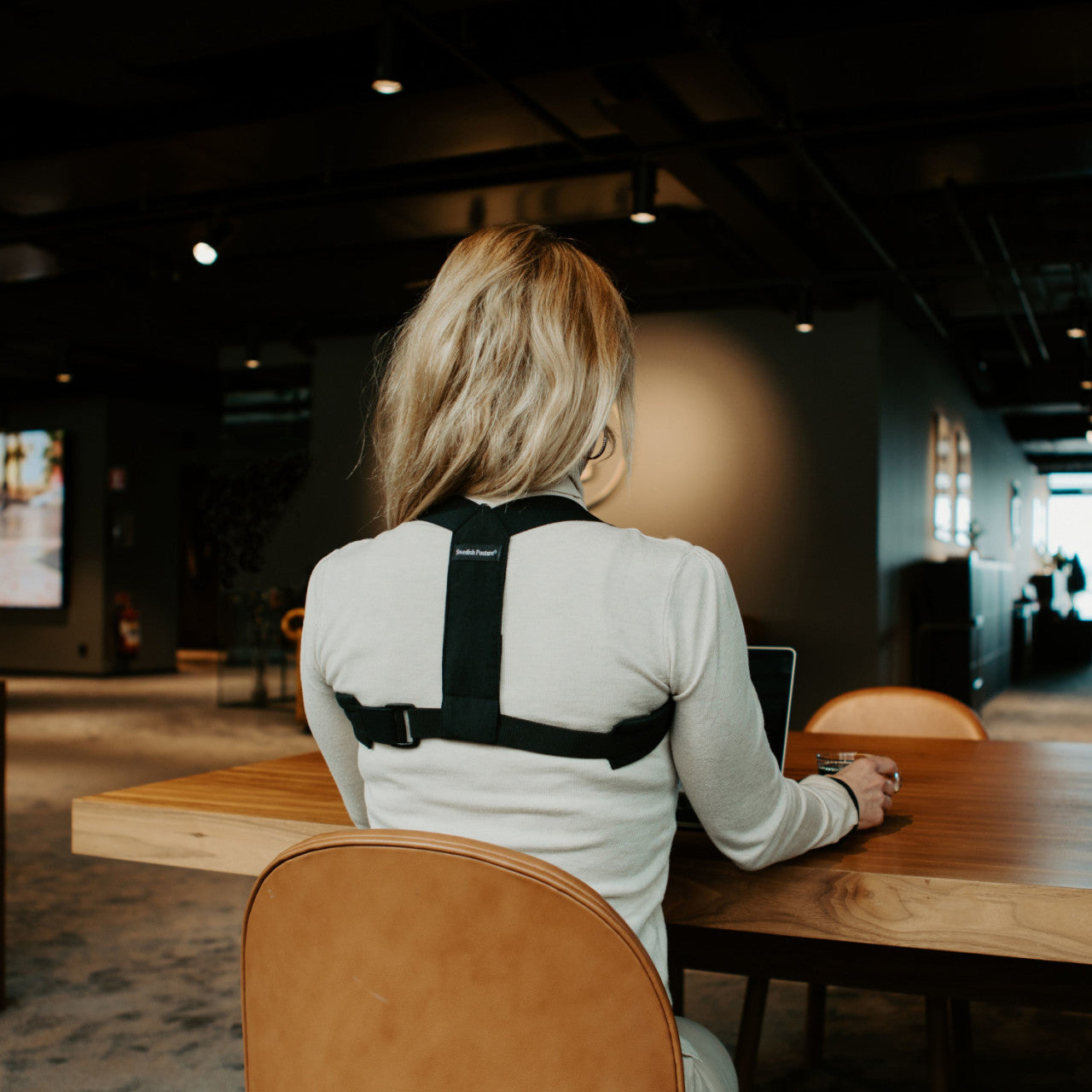 Eine Person mit blonden Haaren trägt eine Haltungskorrektur, während sie an einem Holztisch in einem Büro an einem Laptop arbeitet.