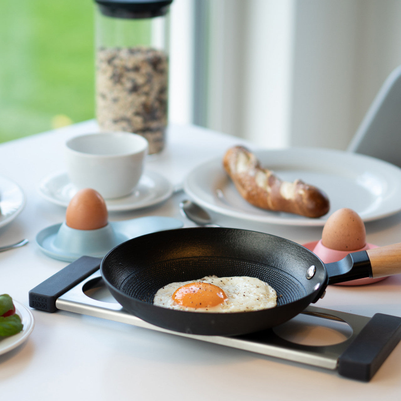 Ein köstliches Spiegelei in einer Pfanne steht auf einem Frühstückstisch mit Eiern, Brot, Müsli und einer Tasse – perfekt zum Genießen mit Ihrer Drip.it Edelstahleinsatz-Abtropfstation für einfaches Aufräumen.