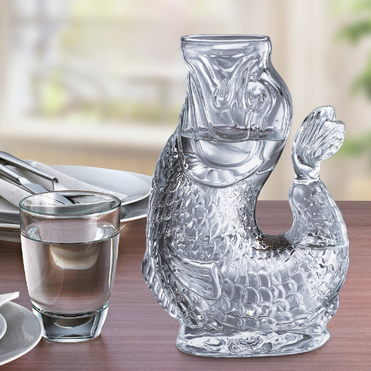 Eine Wasserkaraffe aus Borosilikatglas in Fischform und ein Glas Wasser stehen auf einem gedeckten Holztisch.