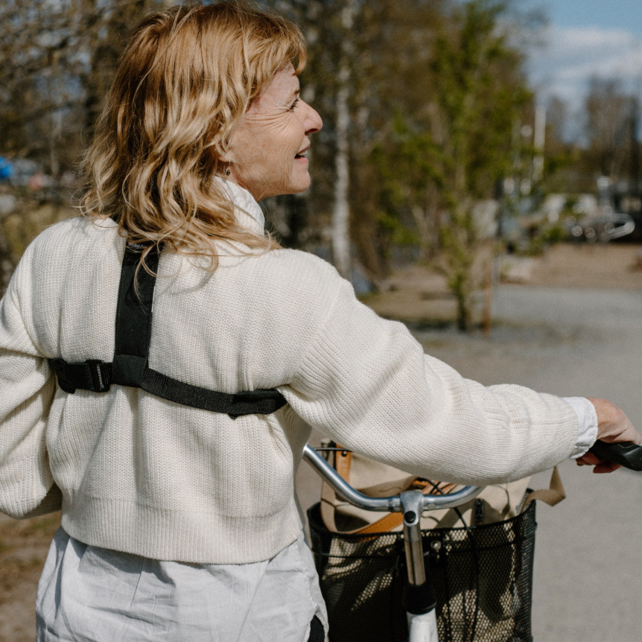 Lächelnde Frau mit Fahrrad geht an einem sonnigen Tag im Freien spazieren, hält den Lenker mit einem Korb davor und genießt die Unterstützung einer Flexi-Haltestütze zur sanften Haltungskorrektur.