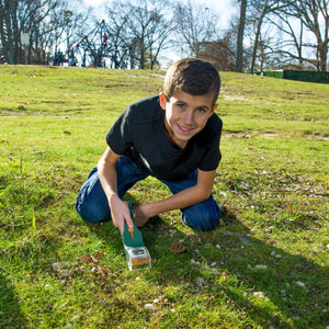 Ein auf Gras kniender Junge untersucht mit einem Vergrößerungsglas den Boden in einem sonnigen Park.