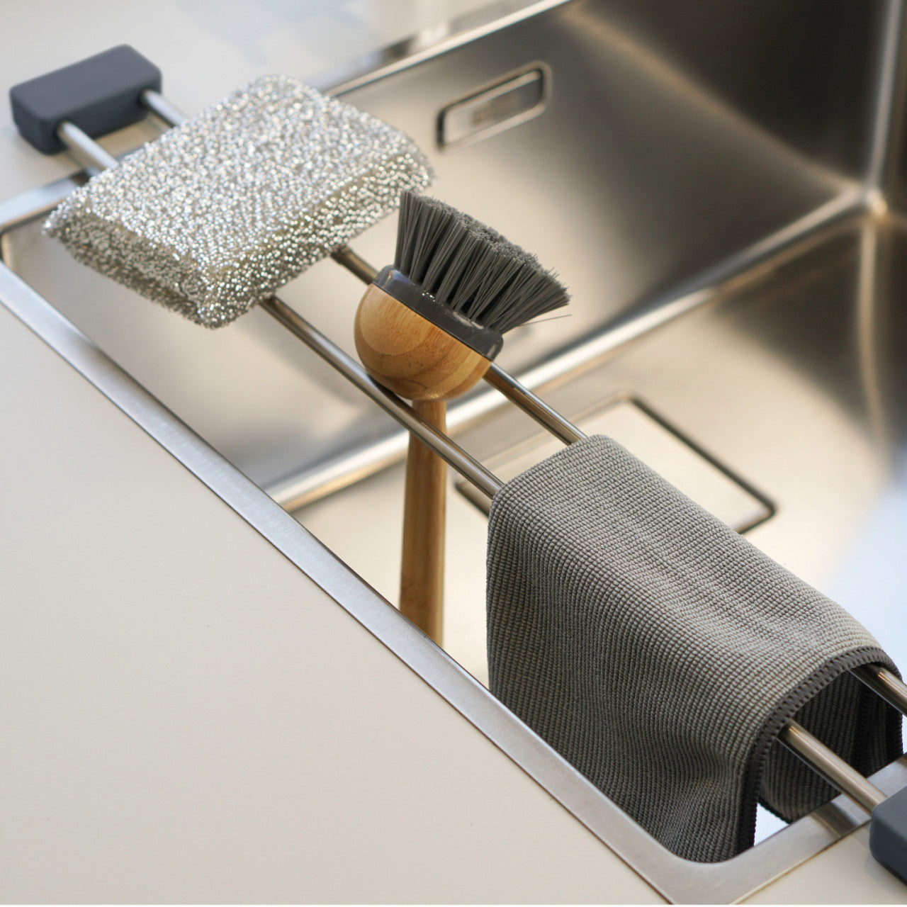Metallspüle mit einer Spültuchhalterstange, an der ein Schwamm, eine Scheuerbürste und ein darüber hängendes graues Tuch hängen – ein unverzichtbarer Küchenhelfer, um Ihren Raum sauber zu halten.
