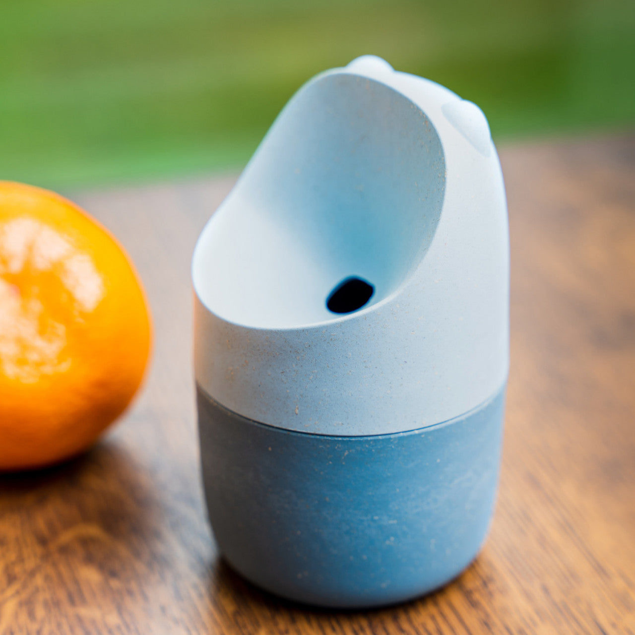 Eine blaue Tasse mit Ausguss aus umweltfreundlichem Holz-Bioverbundstoff steht neben einer Orange auf einem Holztisch mit unscharfem grünem Hintergrund.
