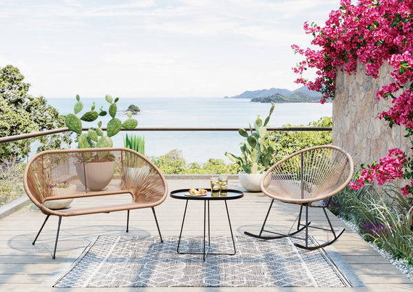 Zwei Korbstühle im eleganten Acapulco-Design und ein kleiner Tisch stehen auf einer Terrasse mit Meerblick, üppigen Pflanzen und leuchtend rosa Blumen.