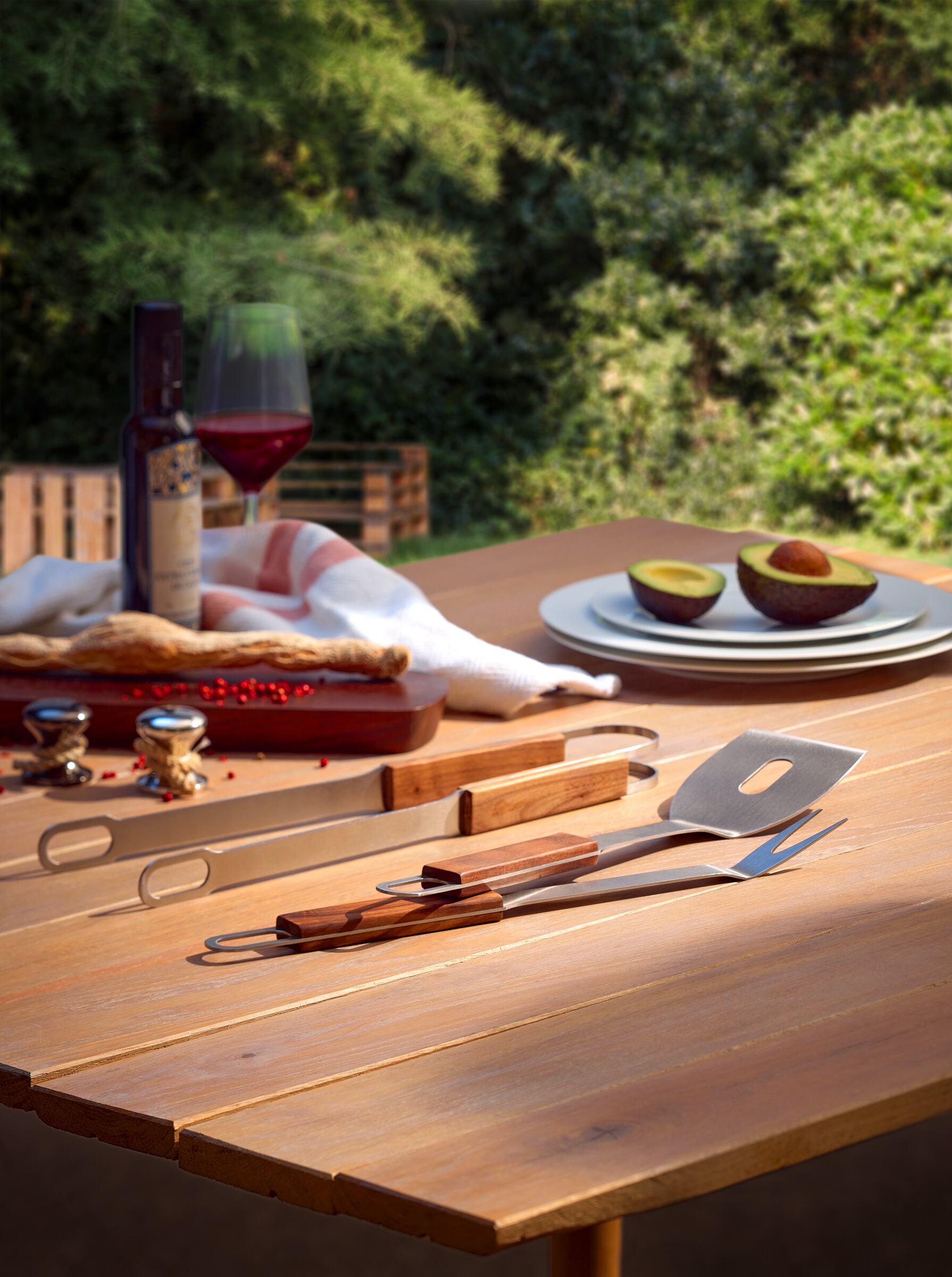 Ein Grillbesteck-Set aus Edelstahl und edlem Walnussholz liegt auf einem Holztisch mit Wein, Tellern, Avocados und grünem Outdoor-Hintergrund.