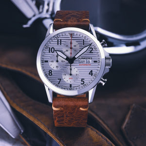 Eine silberne Junkers Aviator Automatik-Armbanduhr mit braunem Lederarmband, Tages- und Datumsanzeige, verfügt über robuste Automatikwerk-Technologie.