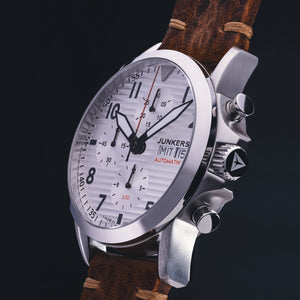 Nahaufnahme einer Junkers Aviator Armbanduhr mit weißem Zifferblatt, Automatikwerk und braunem Lederarmband vor dunklem Hintergrund.
