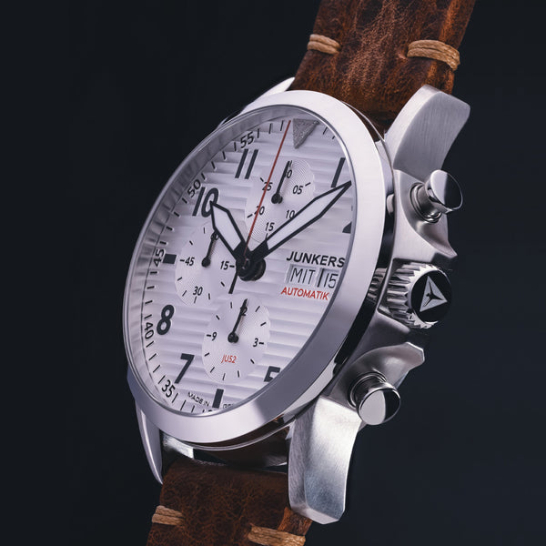 Nahaufnahme einer Junkers Aviator Armbanduhr mit weißem Zifferblatt, Automatikwerk und braunem Lederarmband vor dunklem Hintergrund.