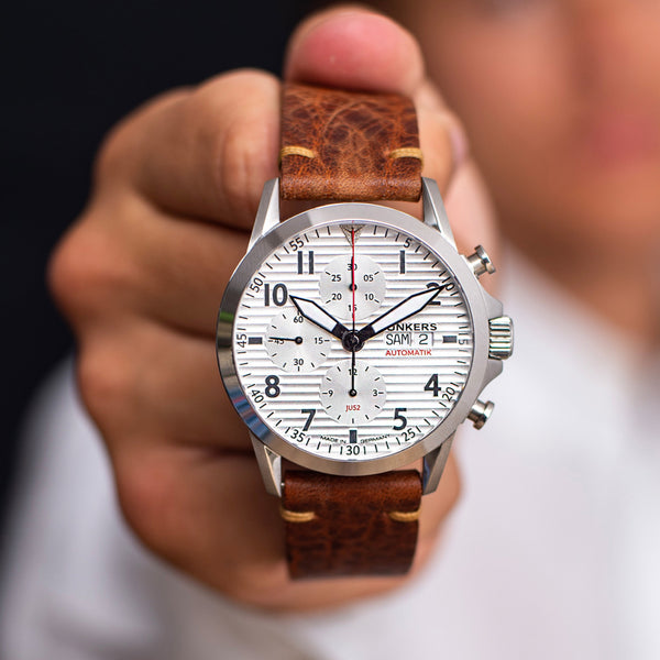 Eine Person hält eine Junkers Aviator Armbanduhr mit Automatikwerk, braunem Lederarmband und weißem Zifferblatt in die Nähe der Kamera.