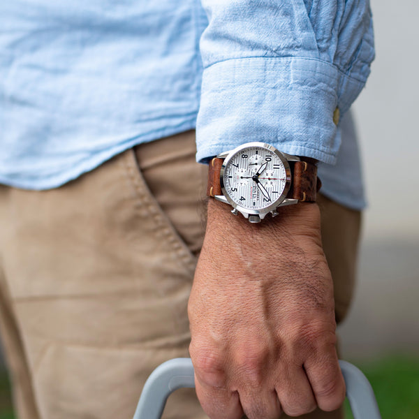Eine Person trägt eine silberne Junkers Aviator Armbanduhr mit Automatikwerk und braunem Lederarmband, dazu ein hellblaues Hemd und hält einen grauen Griff.