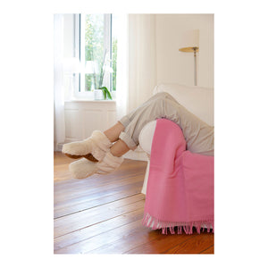 Eine Person lümmelt mit flauschigen Slippies®-Hausschuhen auf einem weißen Sofa, die Beine sind übereinandergeschlagen, und über der Armlehne liegt eine rosa Decke, die für kuscheliger Wärme sorgt.