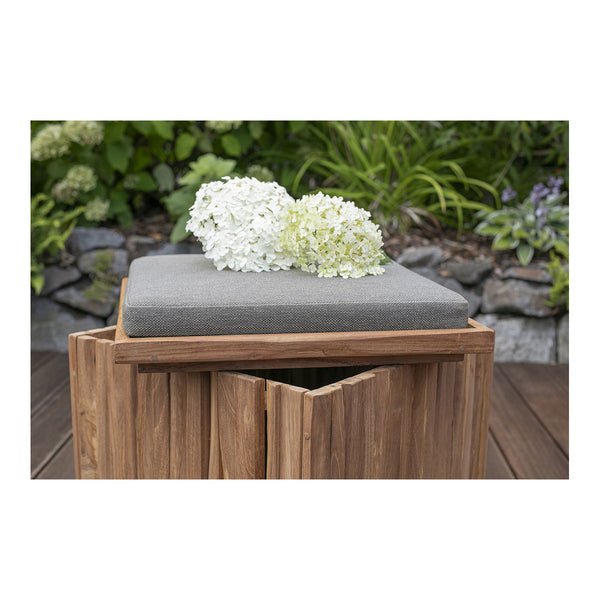 Ein Hocker Oxford aus Teakholz mit einem grauen Kissen und zwei weißen Hortensienblüten darauf, in einem Garten aufgestellt.